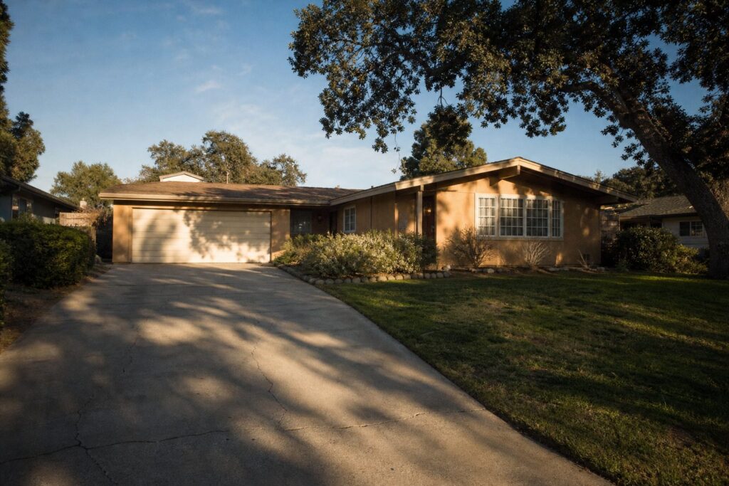 Weathered 1970s ranch home in Valencia with mature oak trees, wide driveway, and dated stucco exterior showing age and character in warm afternoon
